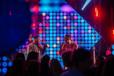 Riga, Letonya - 26 Nisan 2025 - BALTA ikilisi sahnede canlı performans sergiliyor, canlı LED benekli arka plan, her iki şarkıcı da birbirine mikrofonlarla bakıyor, renkli konser enerjisi.