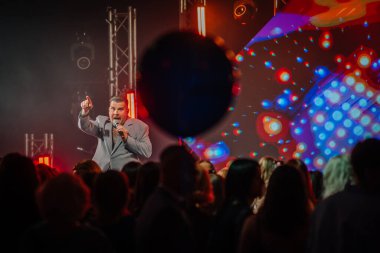 Riga, Letonya - 26 Nisan 2025 - BALTA performans sanatçısı arkasında canlı LED görseller ile canlı bir konser sırasında dramatik sahne aydınlatması altında heyecanlı bir kalabalığa sesleniyor.