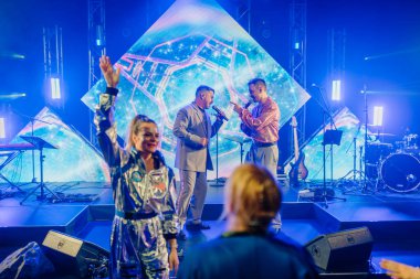 Riga, Letonya - 26 Nisan 2025 - Canlı sahne ile BALTA performansı, LED bilim kurgu arka planı, şarkı söyleyen iki erkek vokal, parlak kostüm sallayan kadın, canlı şovdan keyif alan seyirci.