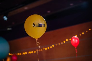 Riga, Letonya - 26 Nisan 2025 - Satürn etiketli sarı bir balon bir BALTA etkinliğinde sergileniyor, etrafları şenlikli tel ışıkları ve diğer renkli balonlarla çevrili.