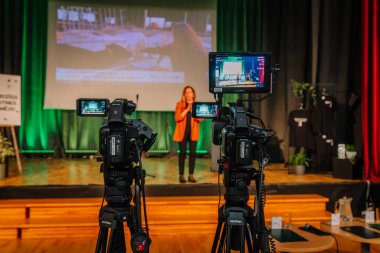 Valmiera, Letonya - 24 Nisan 2025 - Konferans. Profesyonel video kameralar canlı bir etkinlik veya konferansta sahnede bir hoparlör kaydediyor, ekranlar arka planda canlı yayın ve sahne kurulumunu gösteriyor.