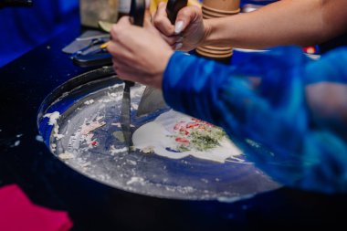 Riga, Letonya - 26 Nisan 2025 - Soğuk tabakta rulo dondurma hazırlayan ellerin yakın çekimi, tatlı tezgahında spatula kullanarak renkli maddeleri harmanlıyor.