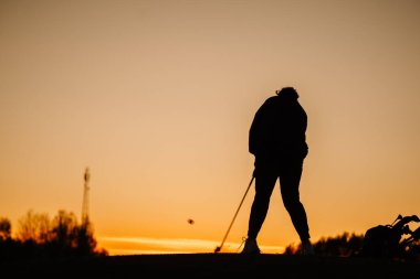 Gün batımında topa vuran bir kadın golfçünün silüeti. Yakınlarda golf çantası ve golf sopası var. Huzurlu bir sahada canlı turuncu bir gökyüzüne çarpıyor..