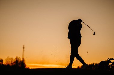 Gün batımında, havada enkaz ve turuncu gökyüzünün altında dinlenen golf çantasıyla güçlü bir takibi tamamlayan bir bayan golfçünün silüeti..