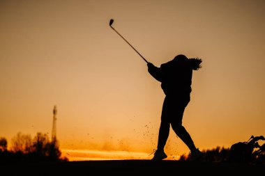Gün batımında dinamik bir salıncak tamamlayan bir kadın golfçünün silüeti. Yanında turuncu bir gökyüzünün altında uçan bir çim ve golf çantası..