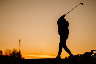 Gün batımında, havada enkaz ve turuncu gökyüzünün altında dinlenen golf çantasıyla güçlü bir takibi tamamlayan bir bayan golfçünün silüeti..