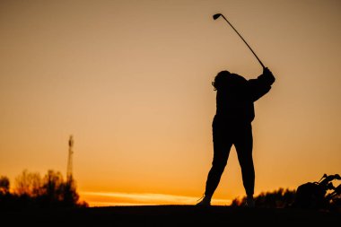 Bir bayan golfçünün silueti gün batımında tam bir salıncak bitiriyor, yanında golf çantası ve arka planda turuncu bir gökyüzü var..
