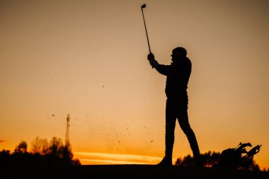 Bir golfçünün silueti gün batımında güçlü bir vuruşu bitiriyor. Yakınlarda uçan çim ve golf çantasıyla, ılık turuncu bir alacakaranlık gökyüzüne karşı..
