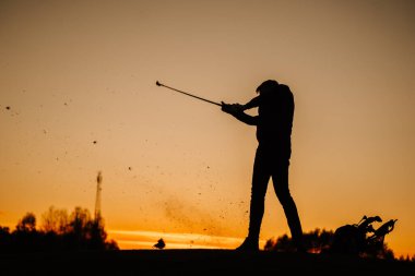 Gün batımında güçlü bir salıncak bitiren bir golfçünün silueti, uçan çimenler ve toprak enkazlarıyla, parlayan turuncu gökyüzü ve yakındaki golf çantasına karşı..