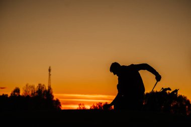 Gün batımında tepede diz çöken bir golfçünün silueti golf çantasıyla çimenlere golf topunu ve golf sopalarını parlayan turuncu gökyüzünün altına yerleştirir..
