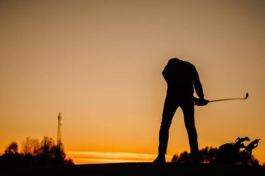 Gün batımında elinde sopayla golf çantasının yanında parlayan turuncu bir gökyüzüne ve uzak ağaçlara bakan bir golfçünün silüeti..