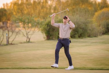 Erkek golfçü bir vuruştan sonra duygusal tepki verir, sopasını kaldırır ve gün batımında bir golf sahasında şapkasına dokunur, ağaçlarla çevrilidir..