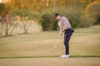 Gün batımında golf sahasında golf oynayan erkek golfçü, topa hassas bir şekilde vurmaya odaklanmış, ağaçlarla çevrili ve açık araziyle çevrili..