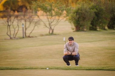 Gülümseyen erkek golfçü yeşil alana çömelmiş, elinde golf sopasıyla atışını değerlendiriyor, manzaralı bir golf sahasında,.