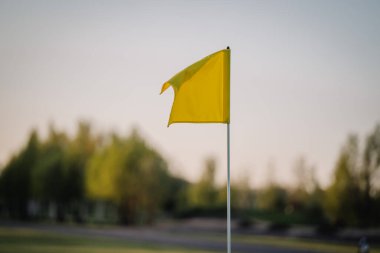 Pistte dalgalanan sarı bir golf bayrağının yakın çekimi, yumuşak doğal ışıklandırma ve arka planda bulanık bir ağaç çizgisi.