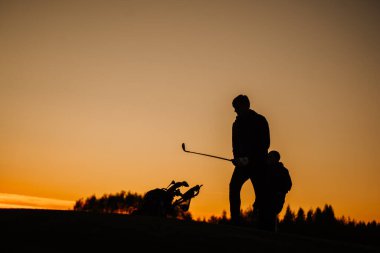 İki golfçü, altın bir günbatımına karşı siluetlerine bürünmüş. Biri elinde golf çantası olan bir sopa tutuyor. Arka planda ağaçlarla dolu huzurlu bir sahada..