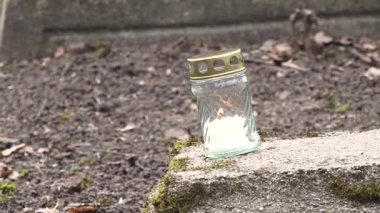 Cesis, Letonya - Ekim 12, 2024 - Metal kapaklı cam bir kavanozda yanan tek bir anıt mum, sessiz, bulutlu bir mezarlıkta yosunlu bir mezara yerleştirildi..