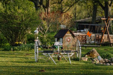 Smiltene, Letonya - 17 Mayıs 2025 Metal bir bahçe dekoru, traktör koltuğu, tekerlekler, asma kaplar ve yemyeşil ve çiçeklerle dolu yemyeşil bir bahçede leylek figürü.