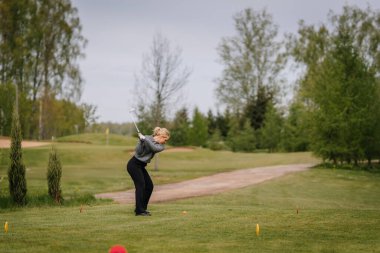 Valmiera, Letonya - 16 Mayıs 2025 - Yeşil pistte kadın golfçü, arka planda ağaçlar ve kum tuzakları ile bulutlu gökyüzünün altında golf oynuyor.