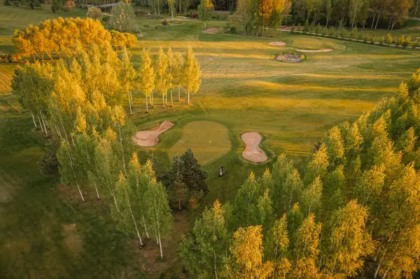 Valmiera, Letonya - 21 Mayıs 2025 - Çevresi altın saatinde canlı ağaçlarla çevrili golf sahasının havadan görünüşü kırsal alana doğru uzanan çim alanları.
