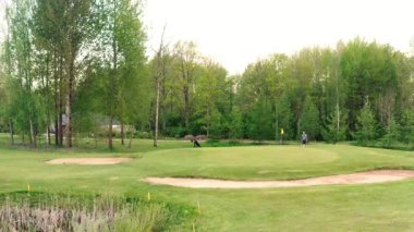 Valmiera, Letonya - 21 Mayıs 2025 - Bir golfçü, uzun ağaçlar ve doğal bitki örtüsüyle çevrili sessiz bir rota çizmeye hazırlanan bir kum sığınağının yakınındaki manikürlü yeşil yolda yürüyor.