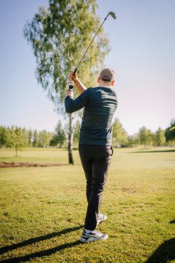 Şapkalı bir adam arka planda ağaçlarla güneşli bir sahada golf sopası sallıyor..