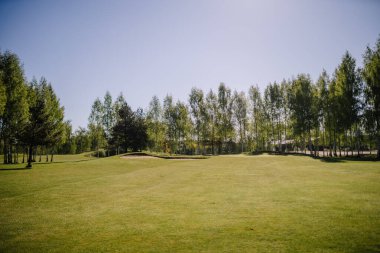 Golf sahasının güneşli manzarası, arka planda kum sığınağı ve ağaçlar var, açık mavi gökyüzünün altında geniş bir kopya alanı var..