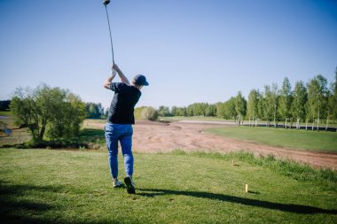 Adam golf sahasından yeşil ağaçlar ve kuru kumlu bir çim yolu bölümüyle çevrili güneşli bir sahada güçlü bir golf sürücüsünü takip ediyor..
