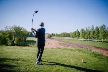 Şapkalı adam, açık mavi gökyüzü ve parlak yeşilliklerle kuru bir çim alanı ve ağaçlarla çevrili bir ufka bakan güneşli bir pistte golf sürücüsünü tamamlıyor..