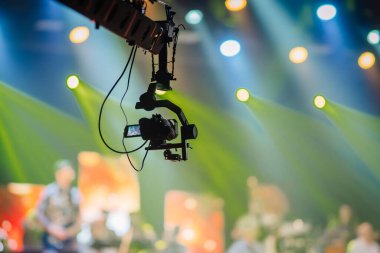 Valmiera, Letonya - 22 Mayıs 2025 - Bir konser veya performans sırasında canlı sahne ışıkları ve renkli bulanık arkaplan ile bir olayı kaydeden askıya alınmış bir kameranın silueti.