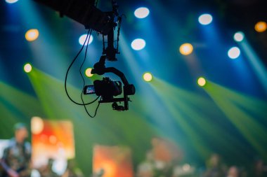 Valmiera, Letonya - 22 Mayıs 2025 - Bir konser veya performans sırasında canlı sahne ışıkları ve renkli bulanık arkaplan ile bir olayı kaydeden askıya alınmış bir kameranın silueti.