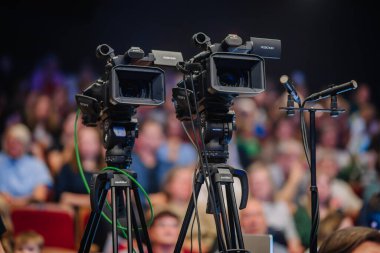Valmiera, Letonya - 22 Mayıs 2025 - İki profesyonel video kamera ve mikrofon kalabalık bir kapalı mekan etkinliğinde kurulmuş, kayıt ya da yayına hazır, arkasında bulanık seyirci ile.