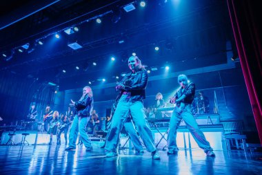 Valmiera, Letonya - 22 Mayıs 2025 - Energetic dans grubu arka planda Yamaha klavyeleriyle parlak bir konser sahnesinde performans sergiliyor. Modern kıyafetler ve senkronize koreografi.