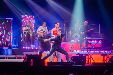 Valmiera, Letonya - 22 Mayıs 2025 - Kırmızılı bir şarkıcının iki erkek dansçı ve renkli konser ışıkları altında canlı Yamaha enstrümanlarının yer aldığı enerjik sahne performansı.