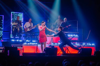 Valmiera, Letonya - 22 Mayıs 2025 - Dinamik sahne performansı kırmızılı bir kadın şarkıcı ve bir erkek dansçı, canlı müzik grubu ve Yamaha klavyesi eşliğinde konser ışıkları altında.