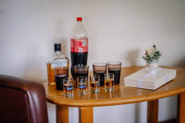 Valmiera, Latvai - 23 Mayıs 2025 - Viski, kola ve shot bardakları bir hediye kutusu ve küçük bir çiçek buketinin yanında ahşap bir masaya dizilmiş, muhtemelen bir düğün tostu için hazırlanmış..