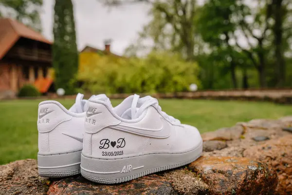 Beyaz Nike Air spor ayakkabıları, baş harfleri ve düğün tarihiyle kişiselleştirilmiş, kırsal taşlara yerleştirilmiş, arka planda bulanık bir bahçe var..