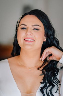 Valmiera, Latvai - 23 Mayıs 2025 - kıvırcık siyah saçlı, inci rengi aksesuarlı, zarif makyajlı, beyaz bir elbise ve altın kolye giyen, düğün gününde evde poz veren gelin.