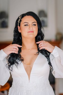 Valmiera, Latvai - 23 Mayıs 2025 - Beyaz elbiseli gelin, kolyesini düzeltiyor, yumuşak makyaj yapıyor ve saç aksesuarı takıyor, düğünden önce kendinden emin ve sakin görünüyor..