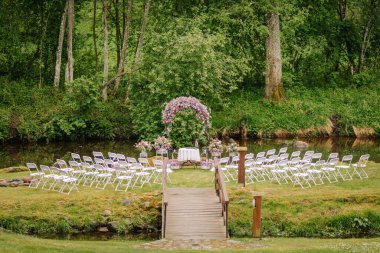 Beyaz sandalyeler, çiçekli kemer ve yemyeşil orman ve dingin doğa ile çevrili bir göl kenarındaki açık hava düğün seremonisi.