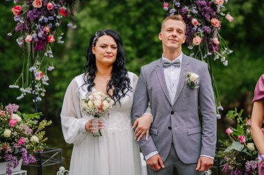 Valmiera, Latvai - 23 Mayıs 2025 - Gelin ve damat, düğün törenlerinde, kol kola, şık giyinmiş ve çiçeklerle çevrili bir şekilde çiçek kemerinin altında dururlar..