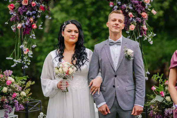 Valmiera, Latvai - 23 Mayıs 2025 - Gelin ve damat, düğün törenlerinde, kol kola, şık giyinmiş ve çiçeklerle çevrili bir şekilde çiçek kemerinin altında dururlar..