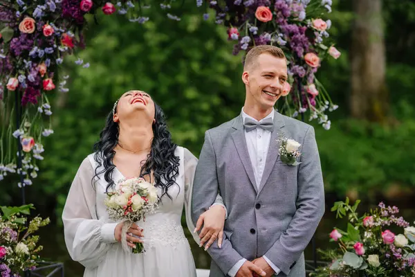 Gelin ve damat düğün törenlerinde çiçek kemerinin altında neşeli bir anı paylaşıyor, yemyeşil bir bahçede gülüyor ve gülüyorlar..