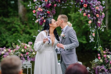 Valmiera, Latvai - 23 Mayıs 2025 - Gelin ve damat, yemyeşil çiçekler ve yeşilliklerle çevrili açık hava düğün töreninde çiçek kemerinin altında şampanya bardakları tutarken öpüşürler..
