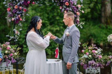 Valmiera, Latvai - 23 Mayıs 2025 - Gelin, çiçek kemerinin altında, canlı çiçek aranjmanları ve yeşilliklerle çevrili bir düğün töreninde damat parmağına yüzük takıyor