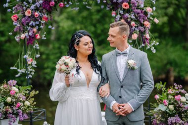 Valmiera, Latvai - 23 Mayıs 2025 - Gelin ve damat, düğün töreninden sonra bir çiçek kemerinin altında, ellerinde buketle birbirlerine bakarak kol kola poz veriyorlar..