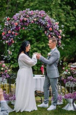 Valmiera, Latvai - 23 Mayıs 2025 - Gelin ve damat, yemyeşil ve renkli çiçek aranjmanlarıyla çevrili bir çiçek kemerinin altında düzenlenen bir açık hava düğün töreninde yüzüklerini değiş tokuş ediyorlar..