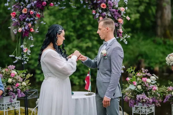 Valmiera, Latvai - 23 Mayıs 2025 - Gelin, çiçek kemerinin altında, canlı çiçek aranjmanları ve yeşilliklerle çevrili bir düğün töreninde damat parmağına yüzük takıyor