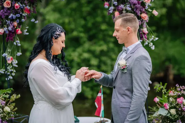 Valmiera, Latvai - 23 Mayıs 2025 - Gelin, hem duygusal hem de dingin görünen çiçek dekoru ve yeşilliklerle çevrili düğün töreninde damatların parmağına yüzük takar.