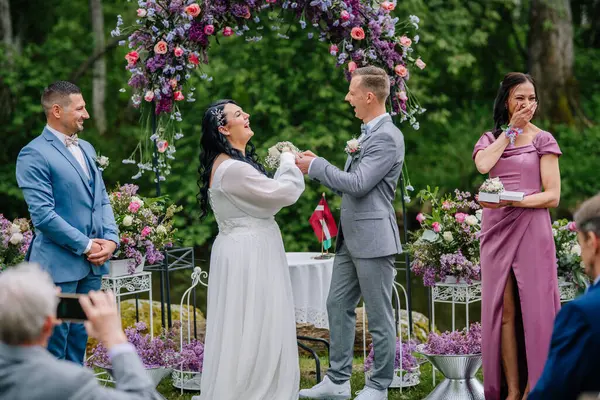 Valmiera, Latvai - 23 Mayıs 2025 - Gelin ve damat olarak neşeli bir an bir açık hava düğünü sırasında yüzüklerinizi takın, etrafında gülümseyen konuklar ve canlı çiçek aranjmanları.